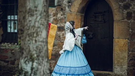 Eine Person in einem historischen Kleid steht vor einer alten Tür. Sie trägt eine Laterne und zeigt in eine Richtung.