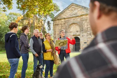 Eine Gruppe von Personen steht im Freien und hört einer Führerin zu, die einen Plan in der Hand hält. Im Hintergrund ist ein historisches Gebäude und Bäume im Herbst zu sehen.