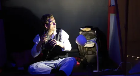 Ein junger Mann sitzt im Dunkeln mit einem Regal und einer Lampe hinter sich. Er hält ein Objekt in der Hand und schaut nachdenklich zur Seite.