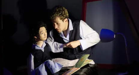 Ein Mann sitzt mit einer Puppe und liest ihr ein Buch vor. Die Szene ist in schwaches Licht getaucht, was eine intime Atmosphäre schafft.