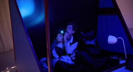 Eine Person sitzt in einem Raum mit gedämpftem, blauem Licht und leuchtet mit einer Taschenlampe nach oben. Neben ihr ist eine Puppe auf dem Bett.