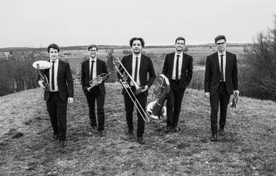 Eine Gruppe von fünf Musikern in Anzügen steht auf einem Hügel. Sie halten verschiedene Blasinstrumente und blicken in die Kamera.