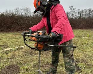 Eine Person mit Schutzkleidung bedient eine Erdbohrer auf einem offenen Feld. Der Himmel ist bewölkt und die Umgebung ist grün mit einigen Hecken im Hintergrund.