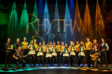 Eine Gruppe von Tänzern in bunten Kostümen steht auf einer Bühne. Im Hintergrund leuchtet das Wort "Rhythm of the Dance" in bunten Lichtern.