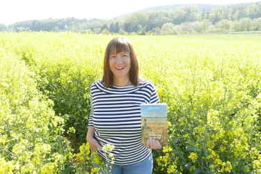 Eine Frau steht in einem gelben Rapsfeld und hält ein Buch in der Hand. Im Hintergrund sind sanfte Hügel und ein klarer Himmel zu sehen.