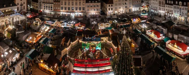 Ein festlicher Weihnachtsmarkt mit vielen bunten Ständen und geschmückten Bäumen. Die Atmosphäre ist lebhaft und einladend, voller Lichter und Menschen.