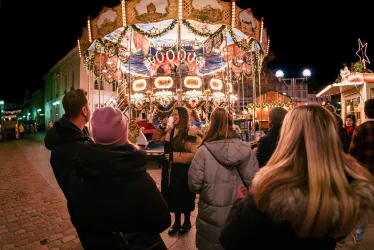 Eine Gruppe von Menschen beobachtet ein festlich beleuchtetes Karussell auf einem Weihnachtsmarkt. Die Atmosphäre ist fröhlich und einladend.