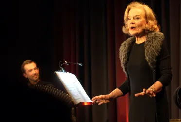 Eine Sängerin steht auf der Bühne und performt, während ein Musiker sie begleitet. Im Hintergrund sind rote und schwarze Vorhänge zu sehen.