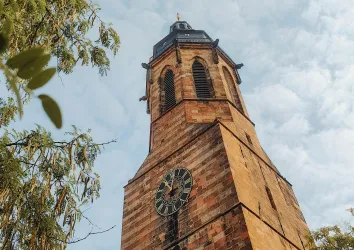 Ein historischer Kirchturm mit einer großen Uhr. Der Himmel ist bedeckt und es sind einige Bäume im Vordergrund sichtbar.