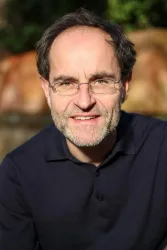 Ein Mann mit Brille lächelt freundlich in die Kamera. Im Hintergrund sind unscharfe grüne Pflanzen zu sehen.