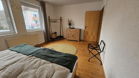 Ein gemütliches Schlafzimmer mit einem großen Bett und Holzfußboden. Der Raum ist hell mit einem Fensterausschnitt und einfachen Möbeln.