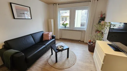 Ein gemütliches Wohnzimmer mit einer schwarzen Couch und einem kleinen Tisch. Der Raum hat große Fenster, die viel Licht hereinlassen, und eine schöne Pflanzendekoration.