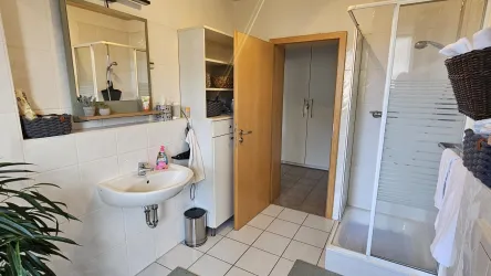 Ein helles Badezimmer mit einem Waschbecken, einer Dusche und einem großen Spiegel. Die Räume sind sauber und modern eingerichtet.