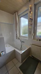 Ein modernes Badezimmer mit einer Badewanne und einem großen Fenster. An der Wand sind dekorative Elemente und ein Teppich auf dem Boden.