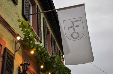 Ein Gebäude mit einem modernen Wappen und einer Fahne. Grünpflanzen schmücken die Fassade und es ist bewölkt.