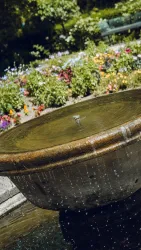 Ein hübscher Brunnen mit sprudelndem Wasser. Im Hintergrund blühen bunte Blumen.
