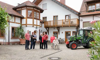 Eine Gruppe von sechs Personen steht vor einem landwirtschaftlichen Gebäude mit einem Traktor in der Nähe. Die Architektur ist traditionell und freundlich gestaltet.
