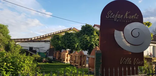 Ein Schild von "Stefan Kuntz" mit dem Text "Villa Vino" ist im Vordergrund zu sehen. Im Hintergrund befinden sich grüne Bäume und ein Gebäude.