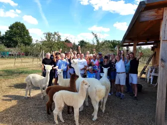 Eine Gruppe von Menschen steht fröhlich zusammen mit mehreren Alpakas auf einem Bauernhof. Im Hintergrund sind Bäume und ein Holzhaus zu sehen.