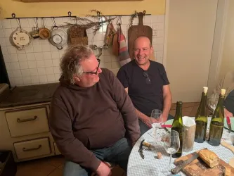 Zwei Männer sitzen an einem Tisch in einer gemütlichen Küche und lachen miteinander. Auf dem Tisch stehen mehrere Weingläser und Flaschen, sowie ein Holzbrett mit Käse.