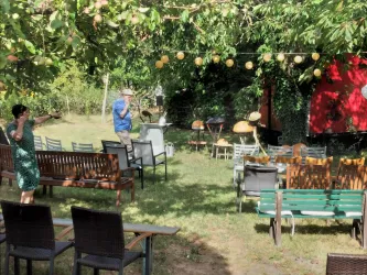 Eine gemütliche Gartenfläche mit mehreren Sitzbänken und Tischen. Über den Sitzbereichen hängen gelbe Früchte, die eine einladende Atmosphäre schaffen.