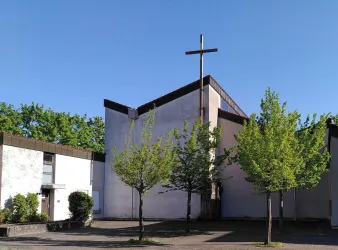 Eine moderne Kirche mit einem Kreuz an der Spitze. Umgeben von grünen Bäumen und klarem Himmel.