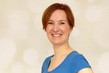 Eine lächelnde Frau mit rotem Haar trägt ein blaues Oberteil. Der Hintergrund ist hell und unscharf, was eine freundliche Atmosphäre schafft.