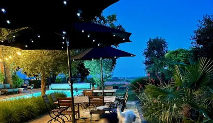 Ein eleganter Außenbereich mit Tischen und Stühlen unter Sonnenschirmen. Im Hintergrund ist ein Pool und üppige Pflanzenlandschaft zu sehen, alles beleuchtet von sanften Lichtern.