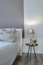 Ein modernes Schlafzimmer mit einem gemütlichen Bett und beigen Kissen. Neben dem Bett steht ein kleiner Beistelltisch mit einer Lampe und einer Pflanze.