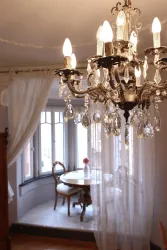 Ein eleganter Kronleuchter hängt von der Decke und beleuchtet einen hellen Raum. Im Hintergrund ist ein runder Tisch mit einem Stuhl zu sehen, der durch Vorhänge von außen getrennt ist.