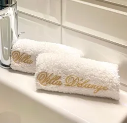Zwei weiße Handtücher mit goldener Schrift "Villa Delange" liegen auf dem Waschbecken. Der Hintergrund ist modern und hell.