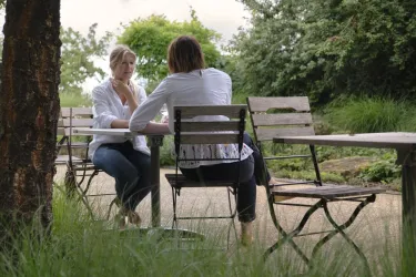 Zwei Frauen sitzen an einem Tisch im Freien und führen ein Gespräch. Sie sind von grüner Umgebung umgeben, mit Bäumen und Gras im Hintergrund.