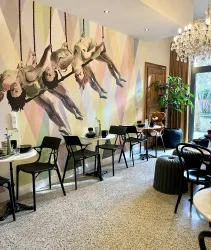 Ein gemütlicher Innenraum mit Tischen und Stühlen. An der Wand befindet sich ein lebendiges Wandgemälde von schwingenden Tänzerinnen.