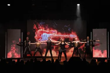 Eine Gruppe von Performern steht auf der Bühne und posiert in einem energetischen Moment. Im Hintergrund ist eine farbige Lichtshow mit feurigen Effekten zu sehen.
