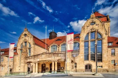 Ein historisches Gebäude mit einer beeindruckenden Fassade und großen Fenstern. Der Himmel ist blau mit einigen Wolken, was für eine schöne Atmosphäre sorgt.