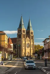 Eine beeindruckende Kirche mit zwei hohen Türmen steht an einer Straße. Die Umgebung ist städtisch und von Bäumen gesäumt.
