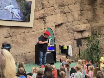 Ein Komiker unterhält eine Gruppe von Kindern auf einer Bühne. Im Hintergrund ist ein Bildschirm mit einem Bild einer weißen Tigerstatue zu sehen.