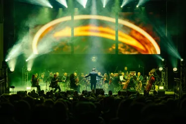 Ein Orchester spielt auf einer Bühne mit bunten Lichtern und einer beeindruckenden Hintergrundprojektion. Der Dirigent leitet die Musiker in einer lebhaften Darbietung.
