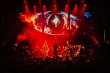 Ein Live-Konzert mit Musikern auf der Bühne und einem beeindruckenden Lichtshow-Hintergrund. Die Atmosphäre ist energiegeladen und das Publikum ist begeistert.