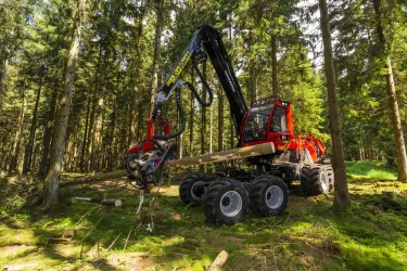 Ein geländegängiger Holzernte-Maschine arbeitet im Wald zwischen hohen Bäumen. Sie hebt einen Holzstamm aus dem Boden.