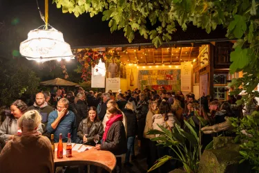 Eine lebhafte Gala im Freien mit vielen Menschen, die an Tischen stehen und sich unterhalten. Die Lichter und die Atmosphäre schaffen eine gesellige Stimmung.