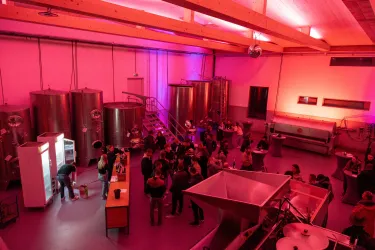 Eine lebhafte Veranstaltung in einer modernen Umgebung mit großen silbernen Tanks. Die Raumbeleuchtung ist in Pink gehalten und viele Menschen genießen die Atmosphäre.