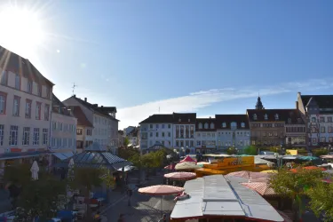 Ein lebhafter Marktplatz mit bunten Ständen und vielen Menschen. Im Hintergrund sind malerische alte Gebäude unter einem klaren blauen Himmel zu sehen.