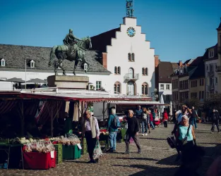 Ein belebter Marktplatz mit Ständen und Menschen. Im Hintergrund steht ein Denkmal auf einem Sockel.