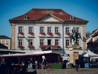 Ein historisches Gebäude mit blühenden Fenstern und einem Reiterstandbild davor. Im Vordergrund sind Marktstände und Menschen zu sehen.