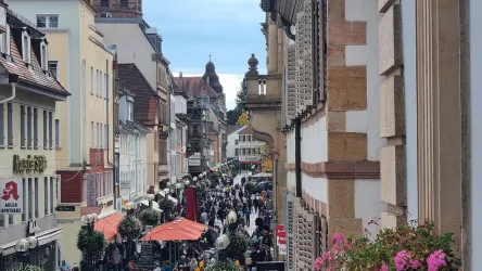 Eine belebte Straße mit vielen Menschen und Tischen im Freien. Im Hintergrund sind historische Gebäude und ein klarer Himmel zu sehen.