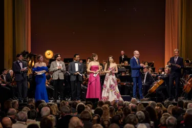 Eine Gruppe von Sängern und Moderatoren steht auf der Bühne vor einem Orchester. Das Publikum sitzt im Vordergrund und schaut gespannt zu.
