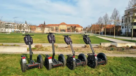 Vier Segways stehen auf einer Wiese vor einem historischen Gebäude. Der Himmel ist teilweise bewölkt und es ist ein klarer Tag.