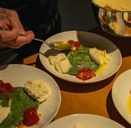 Ein Teller mit grünem Püree, Sahne und Tomatensoße. Eine hand, die mit einem Löffel eine Portion auf den Teller gibt.