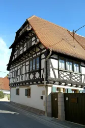 Ein traditionelles Fachwerkhaus mit einem roten Satteldach und großen Fenstern. Das Gebäude steht an einer ruhigen Straße und hat einen grünen Zaun.